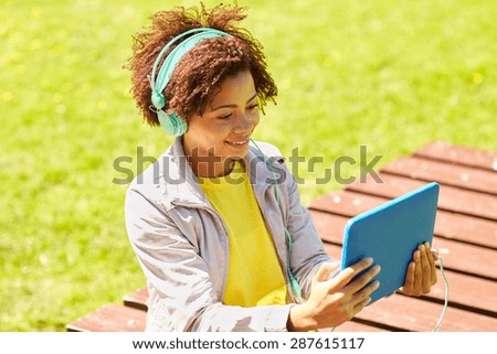 people, summer, technology and leisure concept - happy african american young woman in headphones with tablet pc computer listening to music or watching video outdoors
