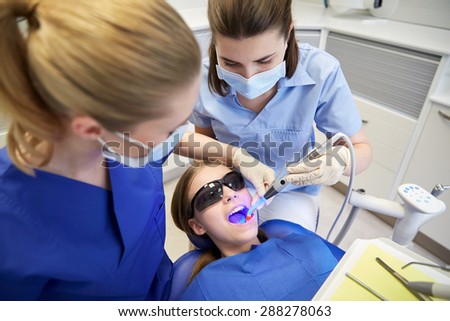 people, medicine, stomatology and health care concept - female dentists with dental curing light and mirror treating patient girl teeth at dental clinic office