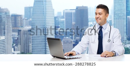 medicine, profession, technology and people concept - smiling male doctor sitting at table with laptop and stethoscope over city background