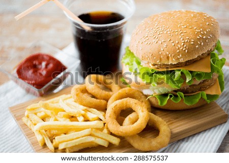 fast food and unhealthy eating concept - close up of hamburger or cheeseburger, deep-fried squid rings, french fries, cola drink and ketchup on wooden table