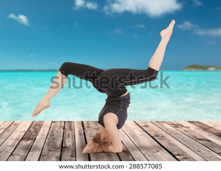 sport, fitness, yoga, people and health concept - happy young woman doing headstand exercise on wooden berth over sea and blue sky background