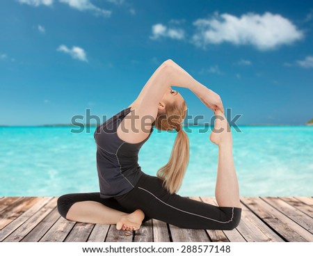people, health, wellness and sport concept - happy young woman in one-legged king pigeon yoga pose on wooden floor over sea and blue sky background