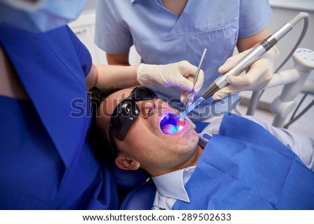 people, medicine, stomatology and health care concept - female dentist and assistant with dental curing light and mirror treating male patient teeth at dental clinic office