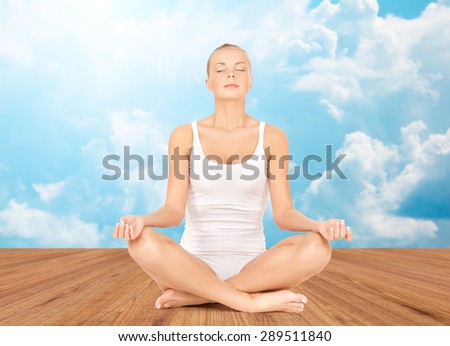 people, health, wellness and meditation concept - woman in underwear meditating in yoga lotus pose on wooden floor over white clouds and blue sky background