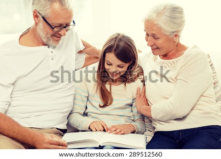 family, generation and people concept - smiling grandfather, granddaughter and grandmother with book sitting on couch at home