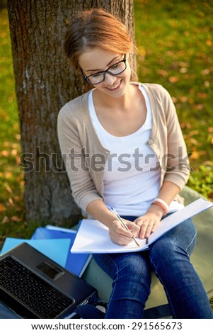 summer, education, learning and teenage concept - happy student girl with laptop computer writing to notebook at campus