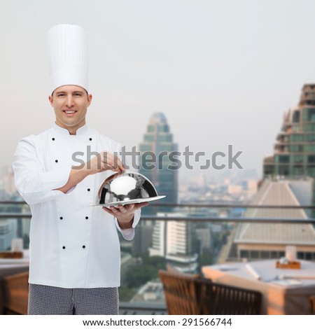 cooking, profession and people concept - happy male chef cook holding cloche over city restaurant lounge background