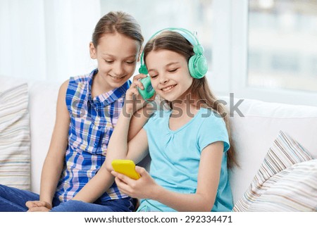 people, children, technology, friends and friendship concept - happy little girls with smartphone and earphones sitting on sofa and listening to music at home