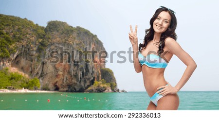 people, gesture, summer, beach and fashion concept - happy woman in bikini swimsuit showing victory hand sign over sea and rock background