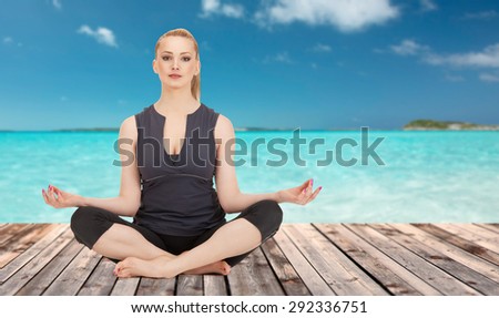 people, health, wellness and meditation concept - happy young woman meditating in yoga lotus pose on wooden floor over sea and blue sky background