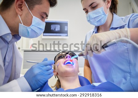 people, medicine, stomatology and health care concept - male dentist and assistant with dental curing light and mirror treating female patient teeth at dental clinic office