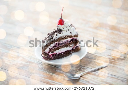 food, junk-food, culinary, baking and holidays concept - piece of delicious cherry chocolate layer cake on saucer with spoon on wooden table over holidays lights background