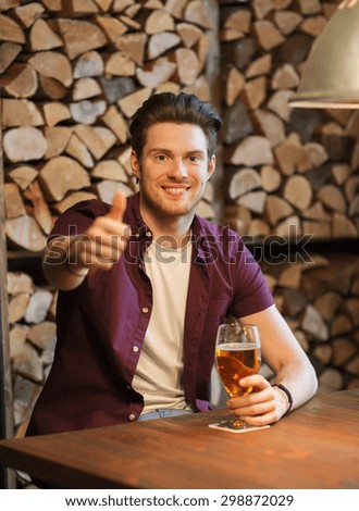 people, drinks, alcohol, gesture and leisure concept - happy young man drinking beer and showing thumbs up at bar or pub