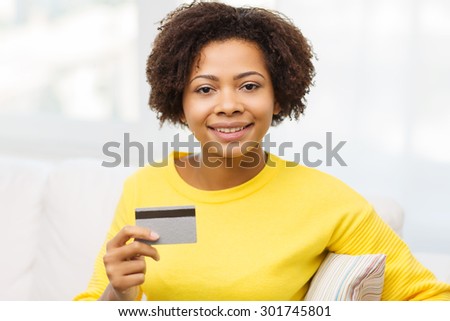 people, bank, shopping and e-money concept - happy african american young woman sitting on sofa with credit or debit card at home