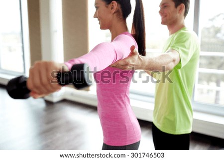 fitness, sport, exercising and people concept - smiling young woman and personal trainer with dumbbells flexing muscles in gym
