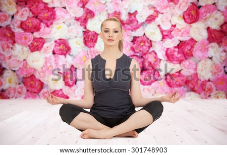 people, health, wellness and meditation concept - happy young woman meditating in yoga lotus pose on wooden floor over wall of flowers background