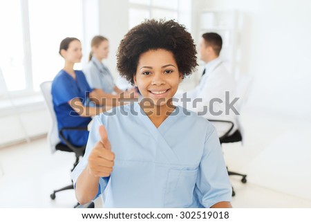 clinic, profession, people and medicine concept - happy  african american female doctor or nurse over group of medics meeting at hospital showing thumbs up gesture