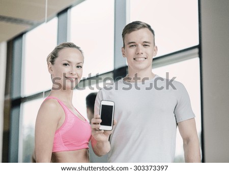 fitness, sport, advertising, technology and diet concept - smiling young woman and personal trainer with smartphone blank screen in gym
