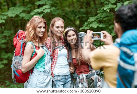 travel, tourism, hike, technology and people concept - group of smiling friends walking with backpacks taking picture by smartphone in woods