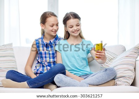 people, children, technology, friends and friendship concept - happy little girls sitting on sofa and taking selfie with smartphone at home