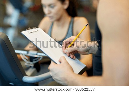 sport, fitness, lifestyle, technology and people concept - close up of trainer hands with clipboard writing and woman working out on exercise bike in gym
