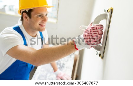 building, profession and people concept - close up of smiling builder in hardhat sanding wall indoors
