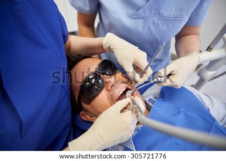 people, medicine, stomatology and health care concept - close up of dentist and assistant hands with dental mirror, drill and air water gun spray treating male patient teeth at dental clinic