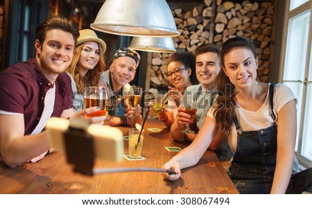 people, leisure, friendship, technology and party concept - group of happy smiling friends with smartphone on selfie stick and drinks taking picture at bar or pub