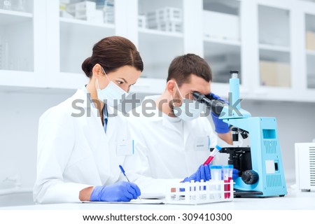 science, chemistry, technology, biology and people concept - young scientists with test tube and microscope making research in clinical laboratory and taking notes