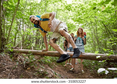 adventure, travel, tourism, hike and people concept - group of smiling friends walking with backpacks and jumping over fallen tree trunk in woods