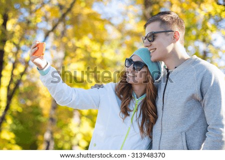 season, vacation, holidays, technology and friendship concept - smiling couple with smartphone taking selfie over autumn park background