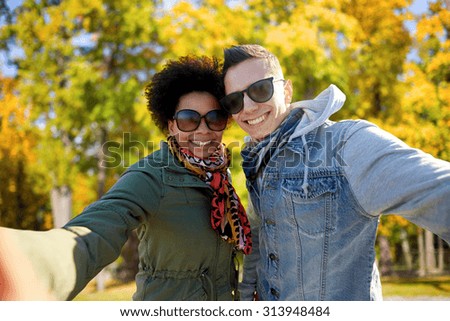 tourism, travel, people, season and technology concept - happy teenage international couple taking selfie over autumn park background