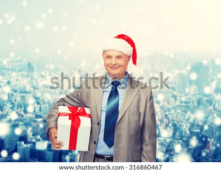 business, christmas, presents and people concept - smiling senior man in suit and santa helper hat with gift over snowy city background