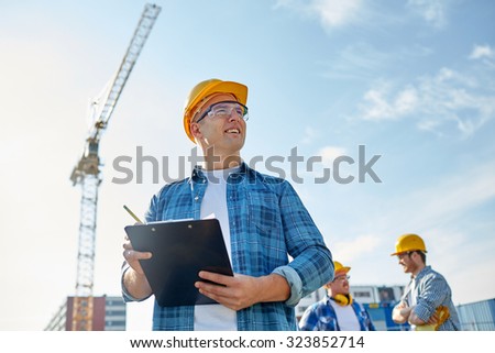 business, building, paperwork and people concept - happy builder in hardhat with clipboard and pencil over group of builders at construction site
