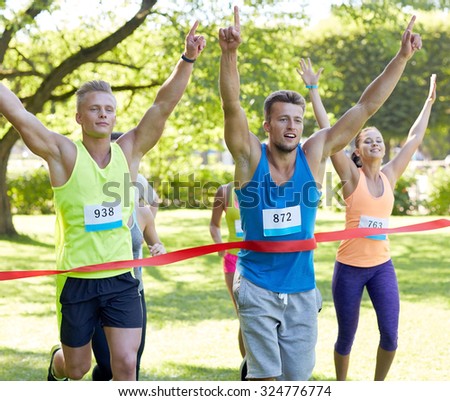 fitness, sport, victory, success and healthy lifestyle concept - happy man winning race and coming first to finish red ribbon over group of sportsmen running marathon with badge numbers outdoors