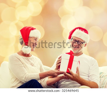 family, holidays, christmas, age and people concept - happy senior couple in santa helper hats with gift box over beige background
