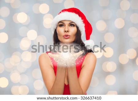 people, holidays, christmas and magic concept - beautiful sexy woman in santa hat and red dress blowing fairy dust off her palms over holidays lights background