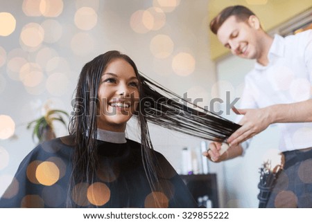 beauty, hairstyle and people concept - happy young woman and hairdresser cutting hair tips at salon over holidays lights