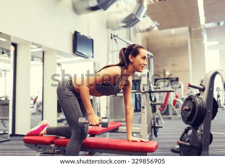 fitness, sport, training and people concept - smiling woman with dumbbell flexing muscles on bench in gym