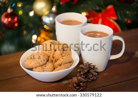 holidays, winter, food and drinks concept - close up of oatmeal cookies, cups with hot chocolate or cocoa drink and pinecones on wooden table over christmas tree background