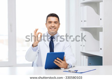 healthcare, profession, people and medicine concept - smiling male doctor in white coat with tablet pc computer showing ok hand sign in medical office