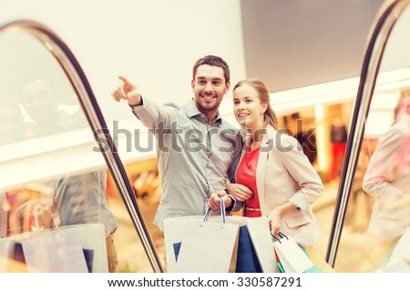 sale, consumerism and people concept - happy young couple with shopping bags rising on escalator and pointing finger in mall
