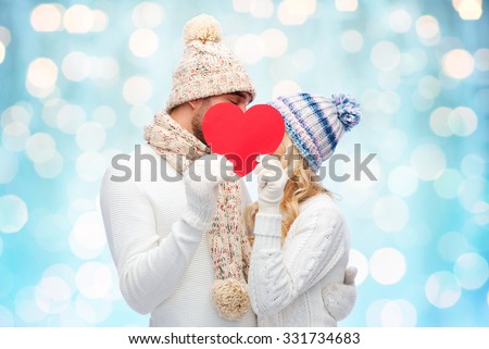 love, valentines day, couple, christmas and people concept - smiling man and woman in winter hats and scarf hiding behind red paper heart shape over blue holidays lights background