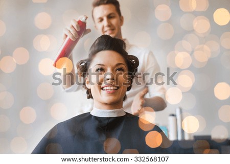 beauty, hairstyle and people concept - happy young woman with hairdresser with hair spray fixating hairdo at salon over holidays lights