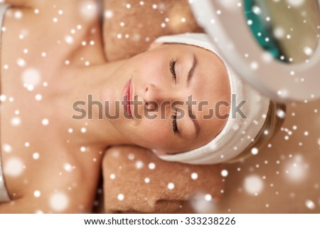 people, beauty, skin care, winter and relaxation concept - close up of beautiful young woman face with closed eyes under magnifying lamp in spa salon with snow effect