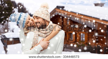 winter, vacation, couple, christmas and people concept - smiling man and woman in hats and scarf hugging over wooden country house and snow background