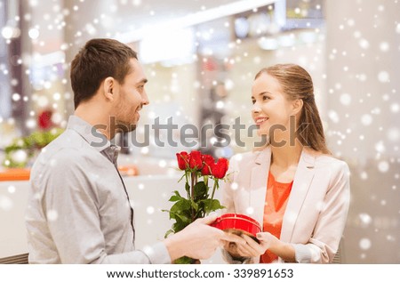 love, romance, valentines day, couple and people concept - happy young man giving red roses and present to smiling woman at cafe in mall with snow effect