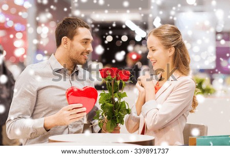 love, romance, valentines day, couple and people concept - happy young man with red flowers giving present to smiling woman at cafe in mall with snow effect