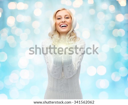 winter, magic, christmas and people concept - smiling young woman in earmuffs and sweater holding fairy dust on palms over blue holidays lights background
