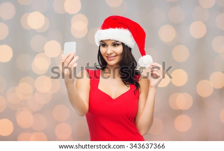 people, holidays, christmas and technology concept - beautiful sexy woman in red santa hat taking selfie picture by smartphone over holidays lights background